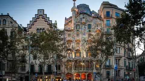 Casa Batlló's mosaic facade and dragon-scale roof lit at dusk on Passeig de Gràcia, with the stepped gable of Casa Amatller beside it and people on the pavement