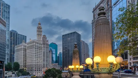 The Chicago River at dusk with tour boats moored below the white Wrigley Building clock tower and lit lamp posts along the river walk
