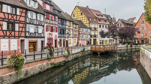 Half-timbered houses in pink, blue and yellow along a canal in Colmar's Little Venice, with flower boxes on the railing and a wooden terrace over the water