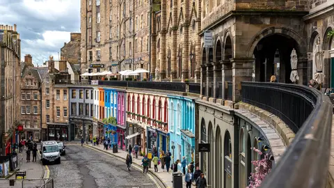 Victoria Street in Edinburgh's Old Town curving downhill past brightly painted shopfronts, with the arched terrace of Victoria Terrace above on the right