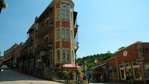 A narrow, wedge-shaped red brick and stone building with iron balconies at the fork of Spring Street and Center Street in downtown Eureka Springs, Arkansas, under a clear blue sky