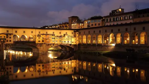 Ponte Vecchio lit up at dusk, its shops and the Vasari Corridor reflected in the still water of the Arno
