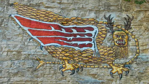 The Piasa Bird painted on the pale limestone bluff above the Great River Road in Alton, Illinois: a scaled, winged creature with antlers, a bearded face and a long tail