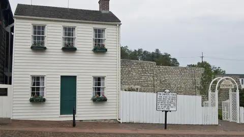 Mark Twain's white clapboard boyhood home in Hannibal, Missouri, with a green door and red flower boxes, beside the whitewashed Tom Sawyer's Fence and its historical marker on a brick street