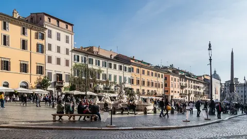 Piazza Navona in Rome on a sunny day, people walking and sitting on benches by the Fountain of Neptune, with the obelisk of the Four Rivers fountain behind