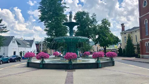 The green bronze Broadway Fountain in Madison, Indiana, ringed by pink flower planters, with brick and painted 19th-century houses behind it under a blue summer sky