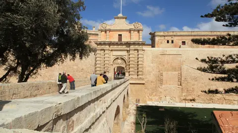Visitors crossing the stone bridge to Mdina's Baroque main gate, with the honey-coloured city walls above a grassy dry ditch and a blue sky
