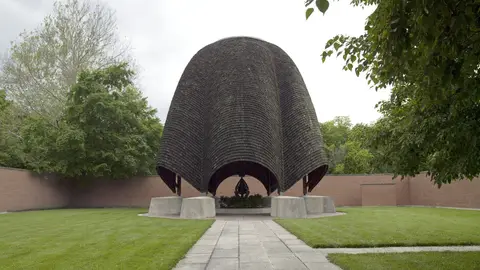 The shingled, bell-shaped dome of the Roofless Church in New Harmony, Indiana, standing over a stone walkway and lawn inside the church's brick walls, with trees around it