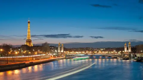 The Eiffel Tower lit gold at dusk on the left, with the Seine and the lamp-lined Pont Alexandre III and its gilded pillars on the right