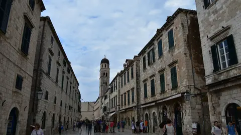 People walking along a stone-paved old town street lined with shuttered limestone buildings, a bell tower at the far end