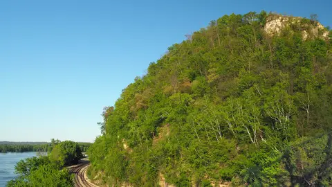 Wooded He Mni Can-Barn Bluff rising above railroad tracks and the Mississippi River in Red Wing, Minnesota, under a clear blue sky