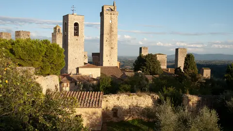 Stone towers of San Gimignano rising above tiled roofs and an old town wall, with olive trees in front and Tuscan hills behind in late afternoon light