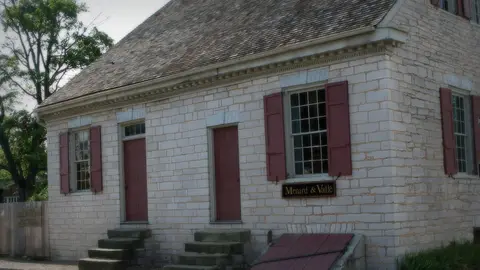 The pale limestone Felix Vallé House in Ste. Genevieve, Missouri, with red shutters, red doors and a Menard & Vallé sign by the street