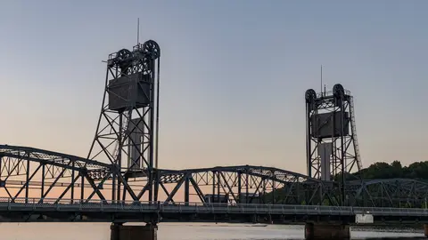 The Stillwater Lift Bridge at dusk, its two steel lift towers and truss spans silhouetted against a pale sky above the calm St. Croix River
