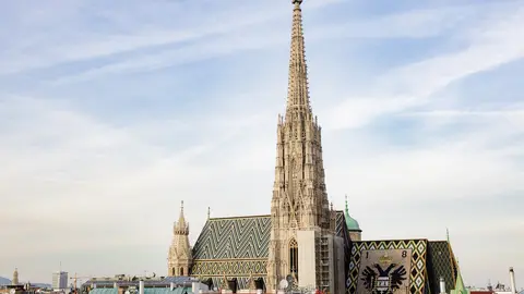 The Gothic South Tower of St. Stephen's Cathedral in Vienna rising above its patterned tile roof and the city's rooftops under a pale blue sky