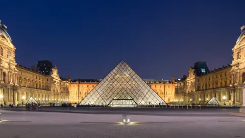 The glass pyramid of the Louvre lit up at dusk in the Cour Napoléon, with the museum's floodlit palace wings on three sides